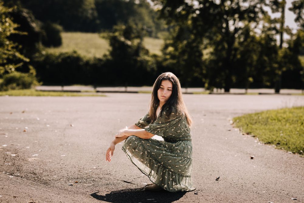 Someone in a patterned green dress crouches on a paved pathway outdoors.