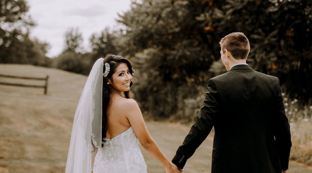 A bride in a white dress looks back while walking hand-in-hand with her groom outdoors.