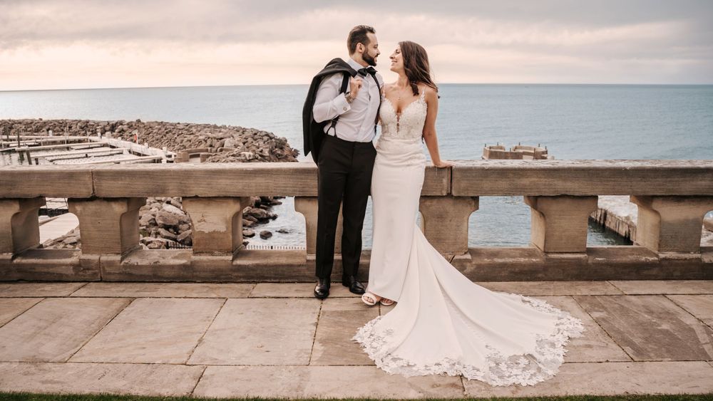Wedding couple poses romantically on oceanfront terrace with marina view at sunset.