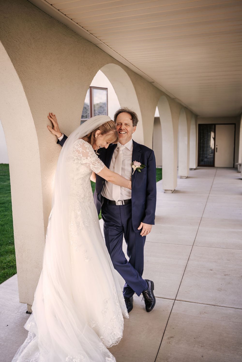A joyful wedding couple shares a candid moment under arched columns in an outdoor corridor.