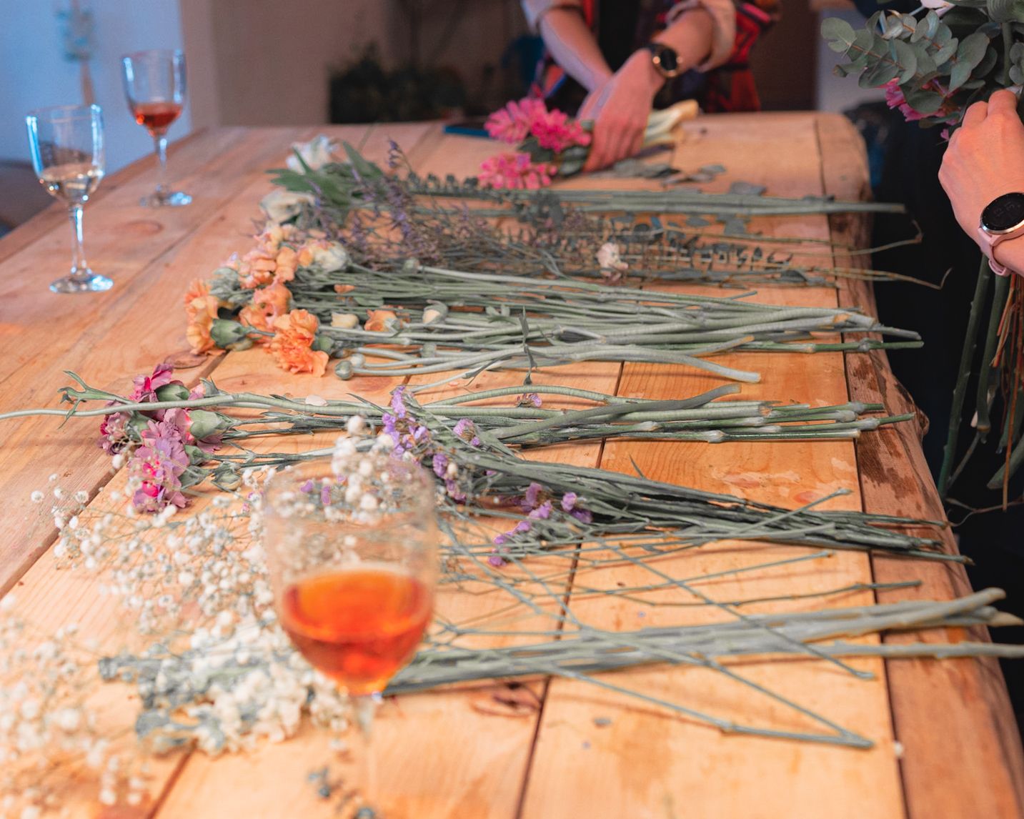 Flower arranging workshop with wine glasses and scattered stems on a wooden table.