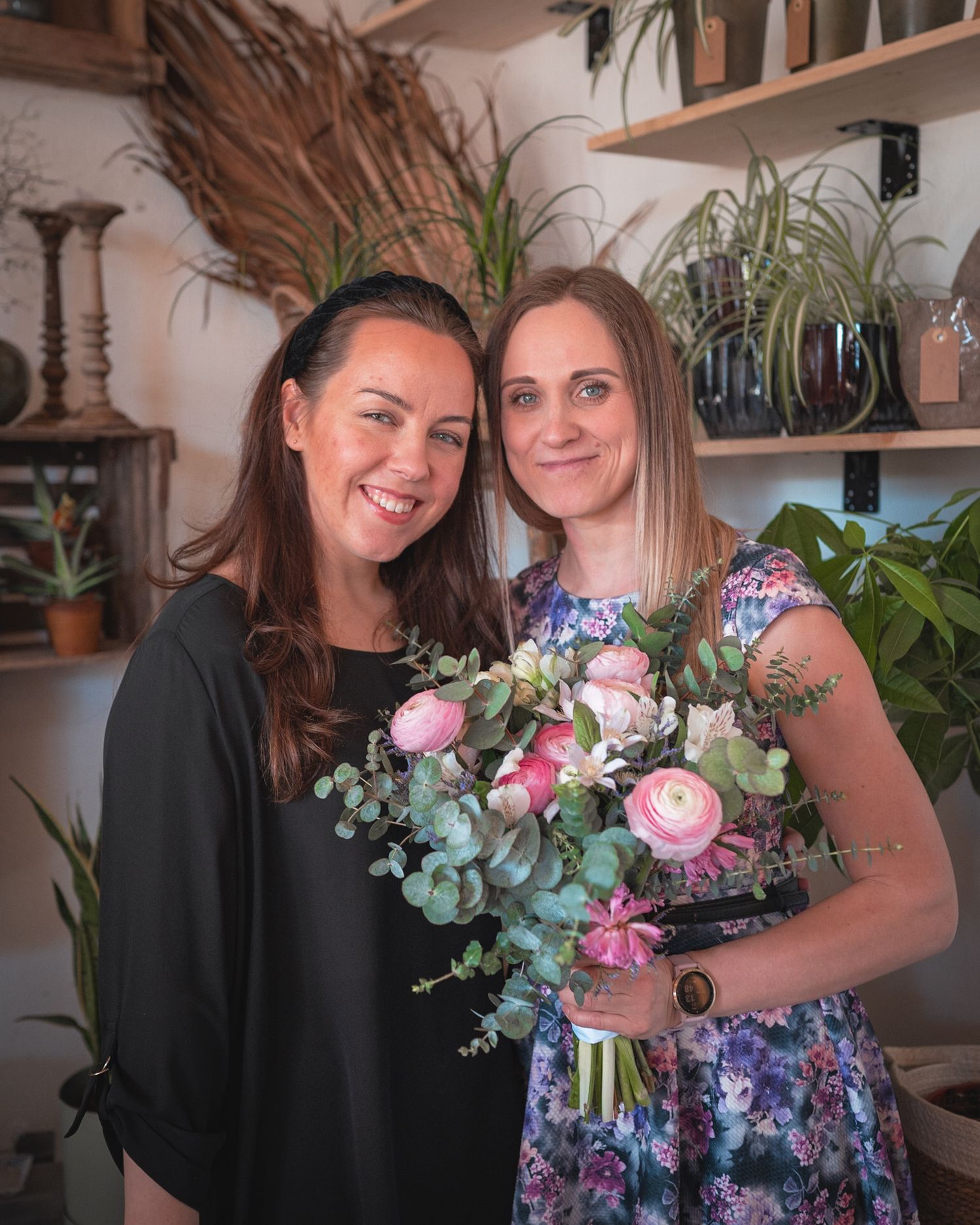 Two people pose with a beautiful floral arrangement in a cozy plant-filled room.