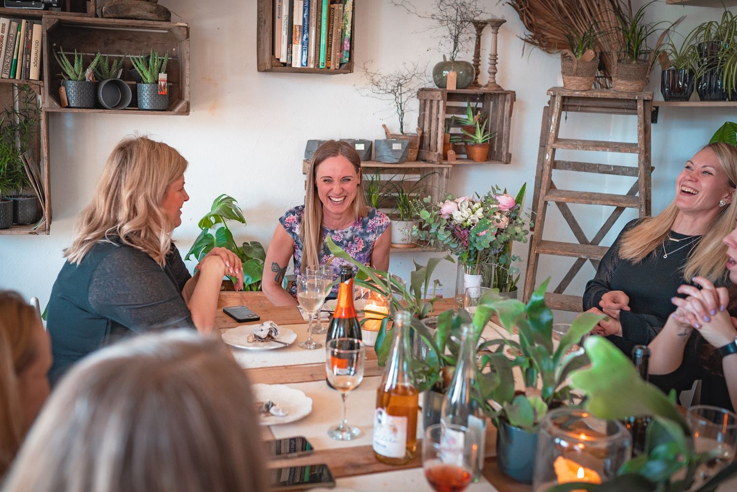 Group enjoying conversation and drinks at a cozy rustic dining space with plants and shelves.