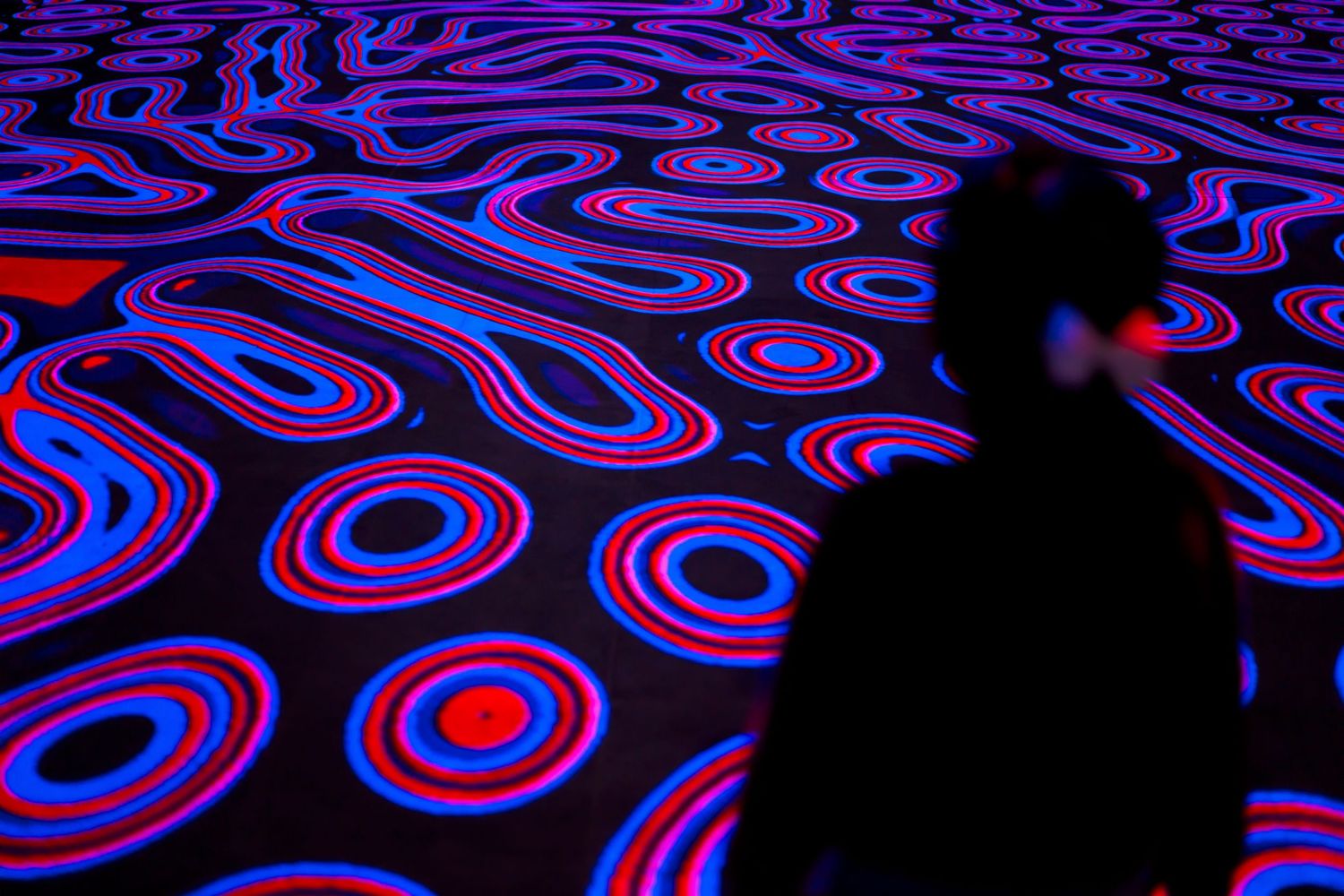 A dark silhouette stands against a vibrant neon floor pattern of concentric circles in blue and red.