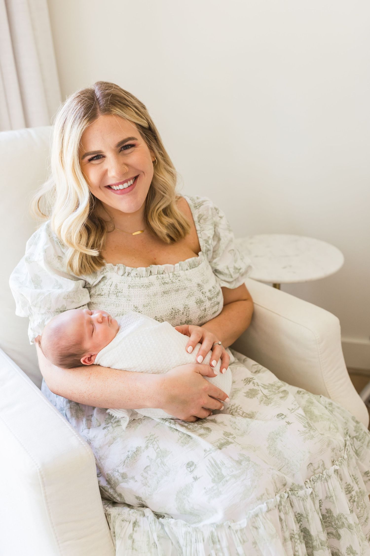 Mother in floral dress holding newborn while sitting in white chair.