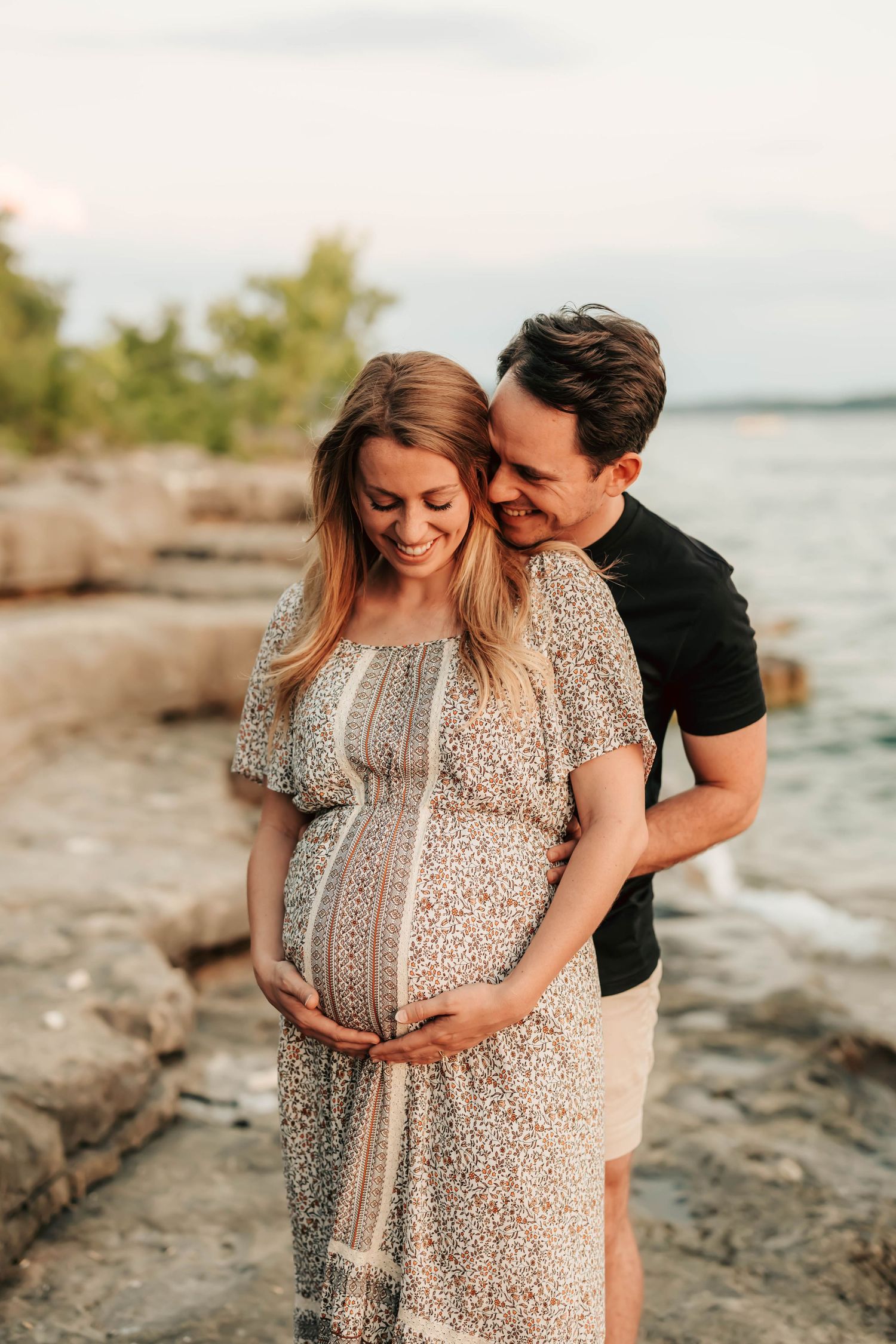 Intimate maternity photo of a couple embracing on a rocky beach at sunset.