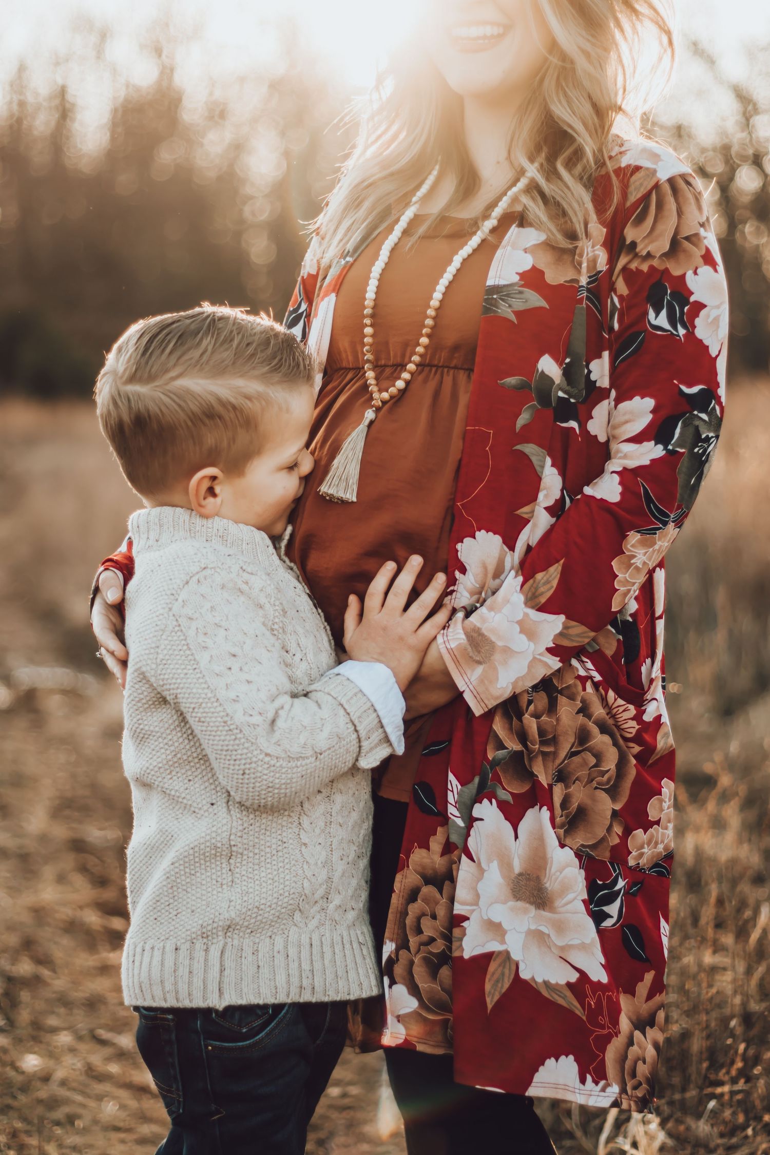 Young child in sweater hugging mother wearing floral dress during sunset photo session.