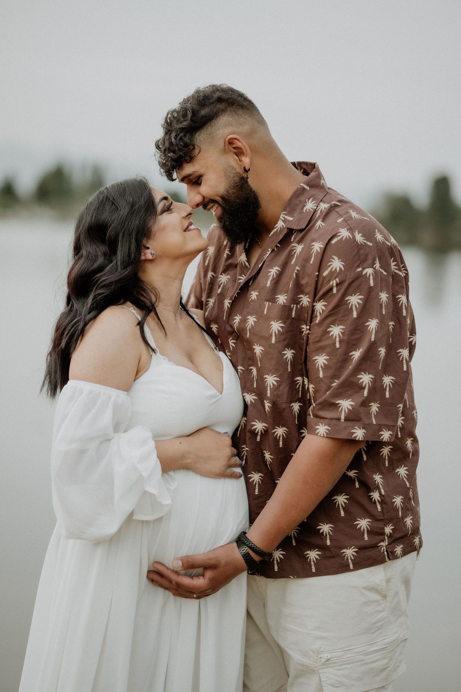Romantic couple in brown and white outfits embracing by the water during sunset.