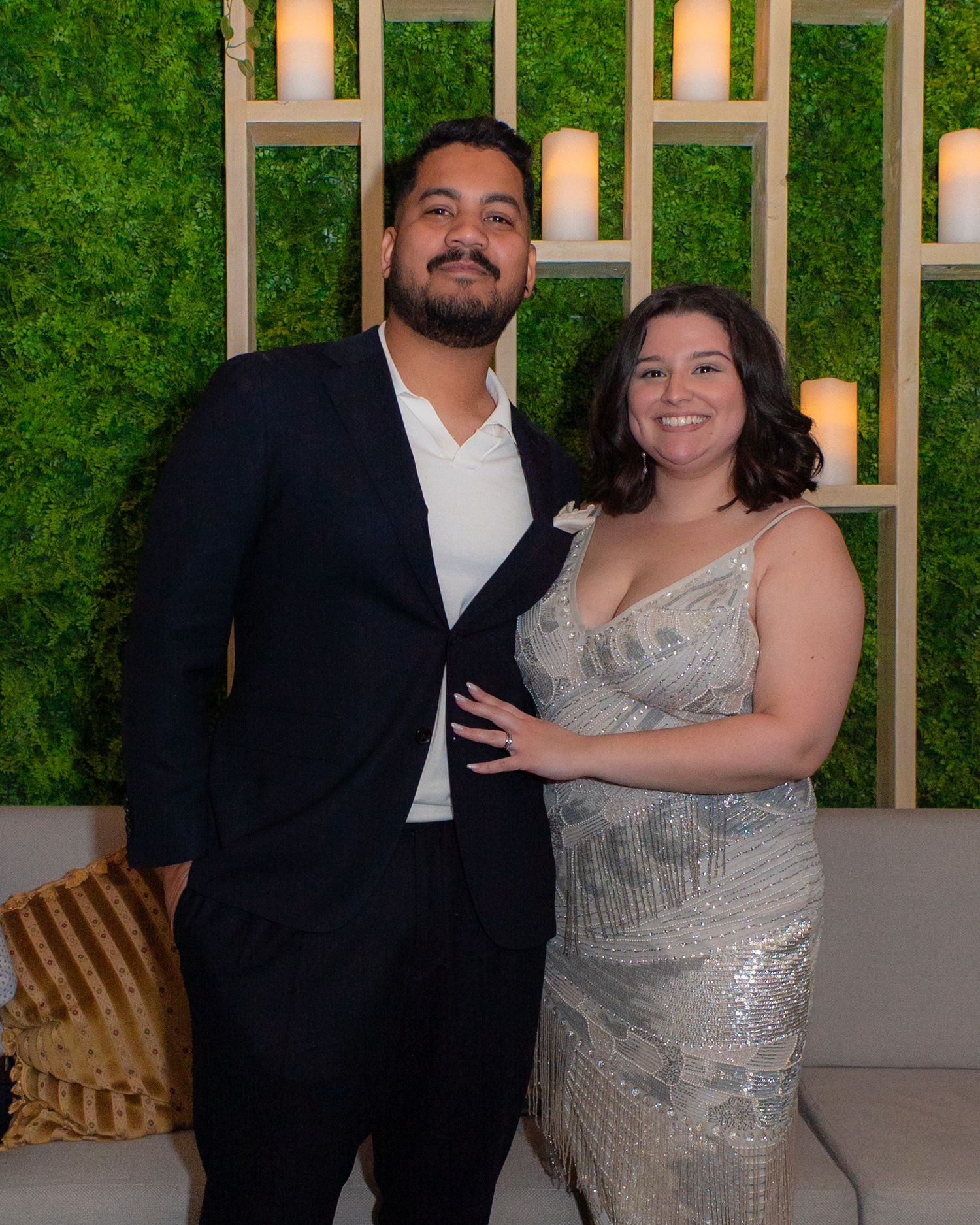 Couple posing together indoors in formal attire in front of green moss floral wall.