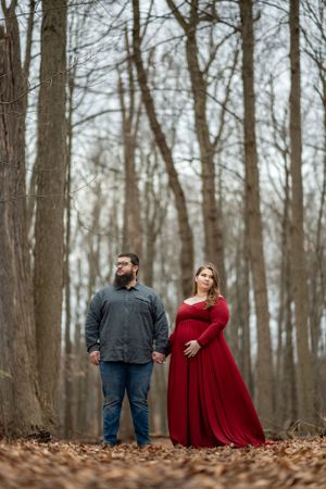 A couple poses in a winter forest scene with bare trees and fallen leaves.