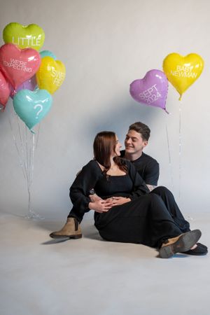 A couple poses with colorful heart-shaped balloons against a grey backdrop.