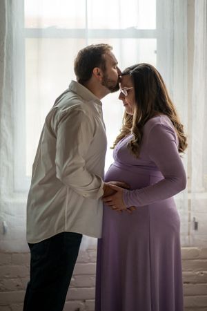 A couple in white and purple embrace near a bright window.