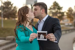 A couple shares a romantic moment while holding an ultrasound photo outdoors.