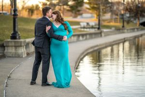 A couple embraces by a lakeside path at sunset, with the woman wearing a flowing turquoise gown.