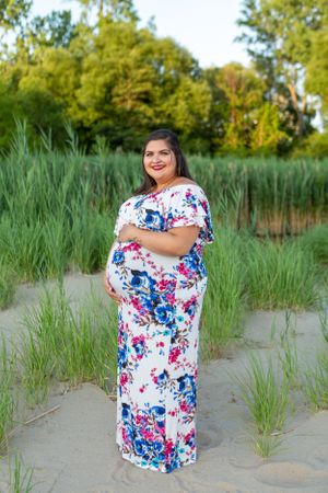 A maternity portrait of someone wearing a floral maxi dress standing among beach grass at golden hour.