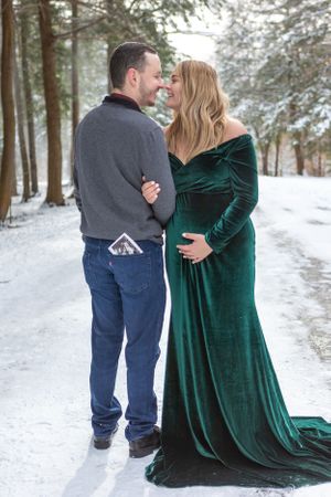 A winter maternity photo featuring a luxurious green velvet dress against a snowy forest backdrop.