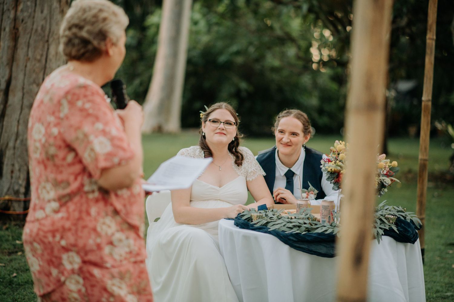 Intimate outdoor wedding celebration with couple seated at decorated table beneath tall trees.