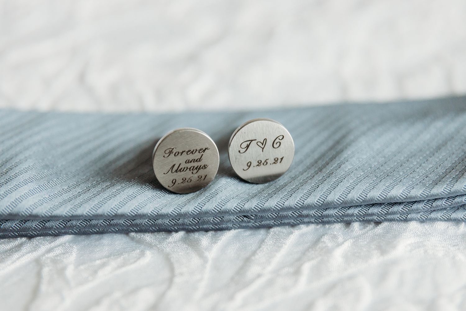 Two engraved silver cufflinks with initials and dates displayed on light blue fabric.