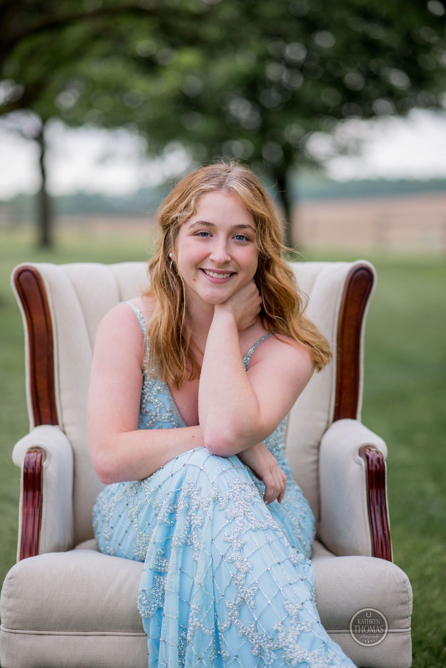 Portrait of a woman in a light blue formal dress sitting in a cream colored wingback chair outdoors.
