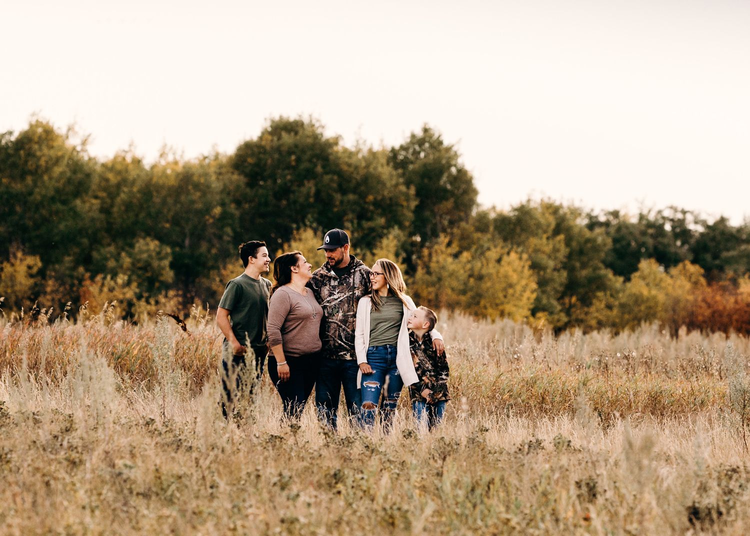 Family of five standing together in a golden autumn field surrounded by colorful fall foliage.