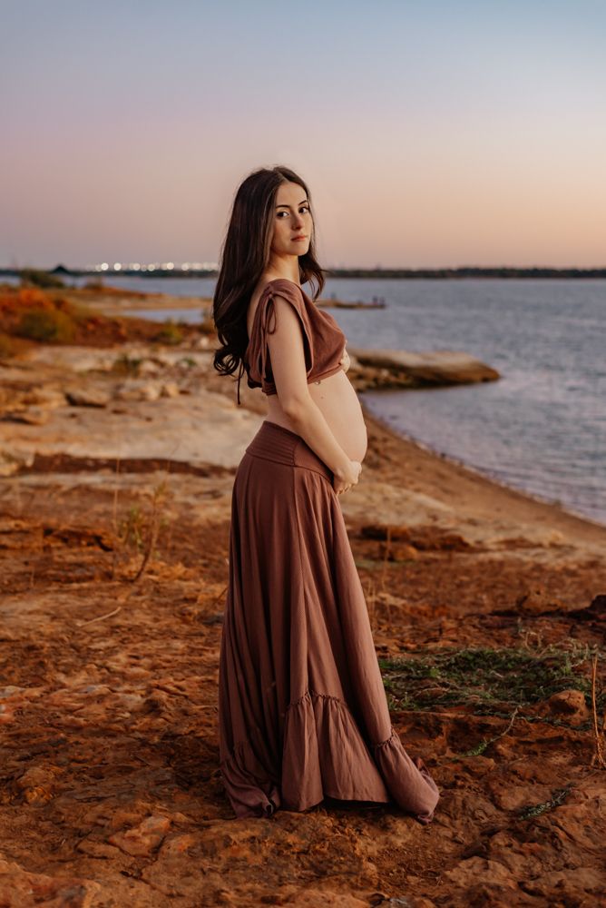 Maternity photo by the water with burgundy dress against sunset sky and rocky shore.