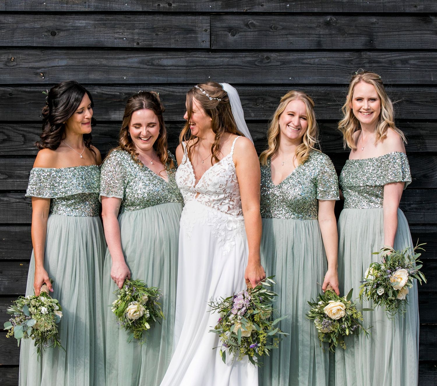 Wedding party in sage green dresses holding natural bouquets against dark wooden wall.