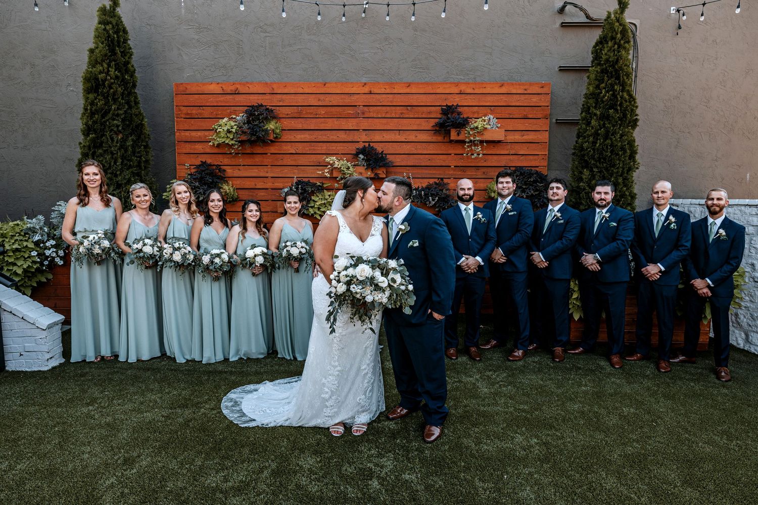 A wedding party poses against a wooden wall with bridesmaids in sage green and groomsmen in navy suits.