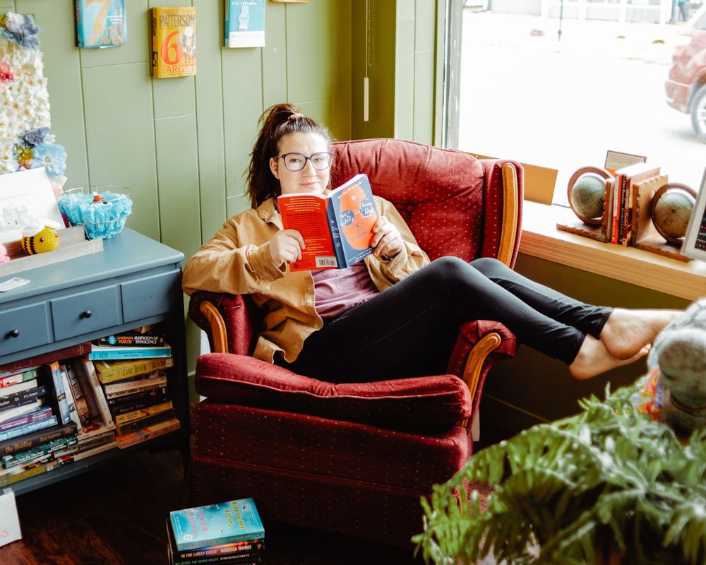 A cozy reading scene in a home library with someone lounging in a red armchair with a dog and surrounded by books.