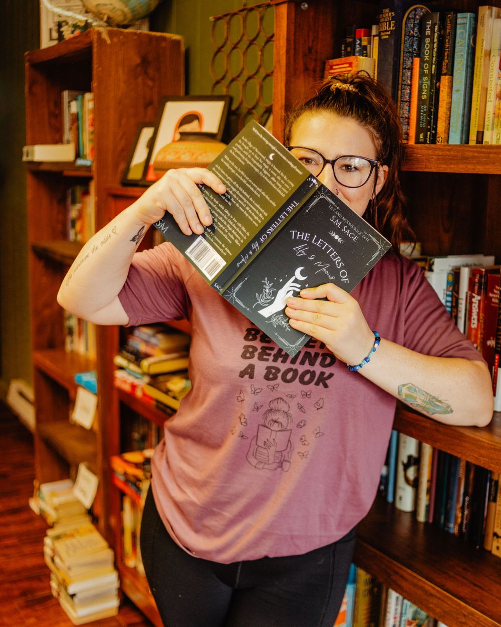 Person in glasses holding books in front of wooden bookshelf wearing pink shirt with book-themed graphic.