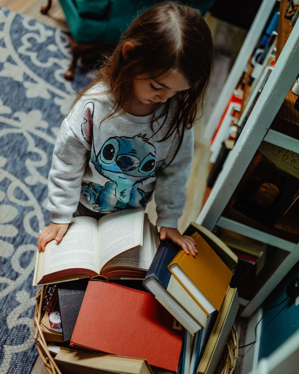 Child wearing Stitch sweatshirt reading book while surrounded by stacks of books on patterned rug.