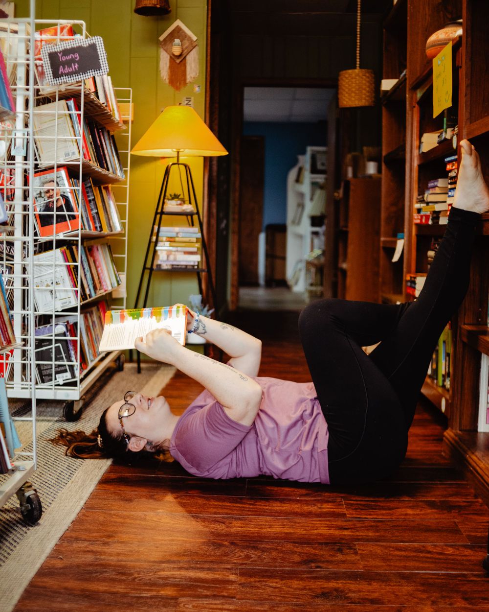 Someone lying on wooden floor reading book next to bookshelf and yellow lamp in bookstore.