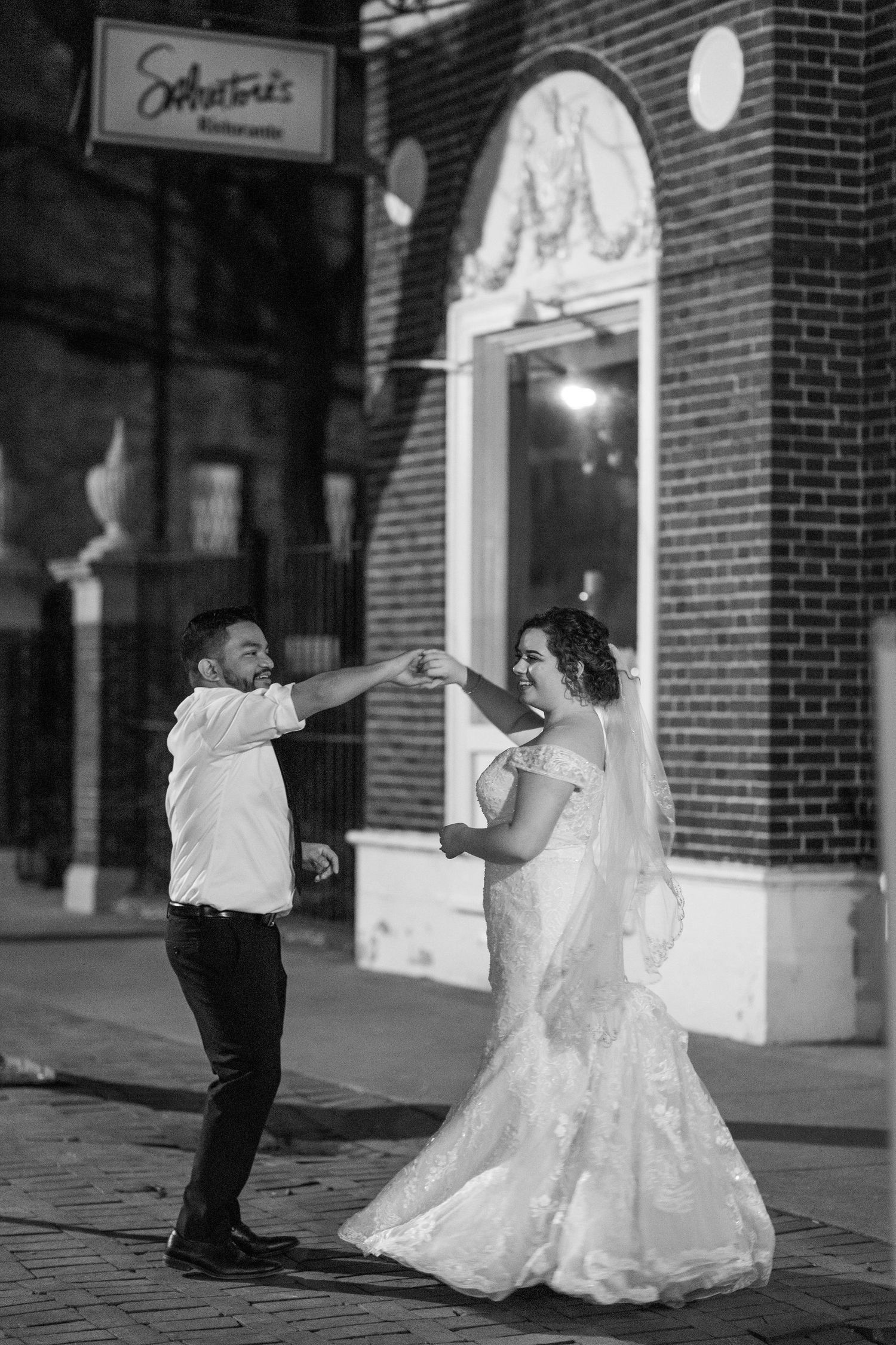 A couple dancing and laughing together on a street at night outside a brick building with vintage signage.