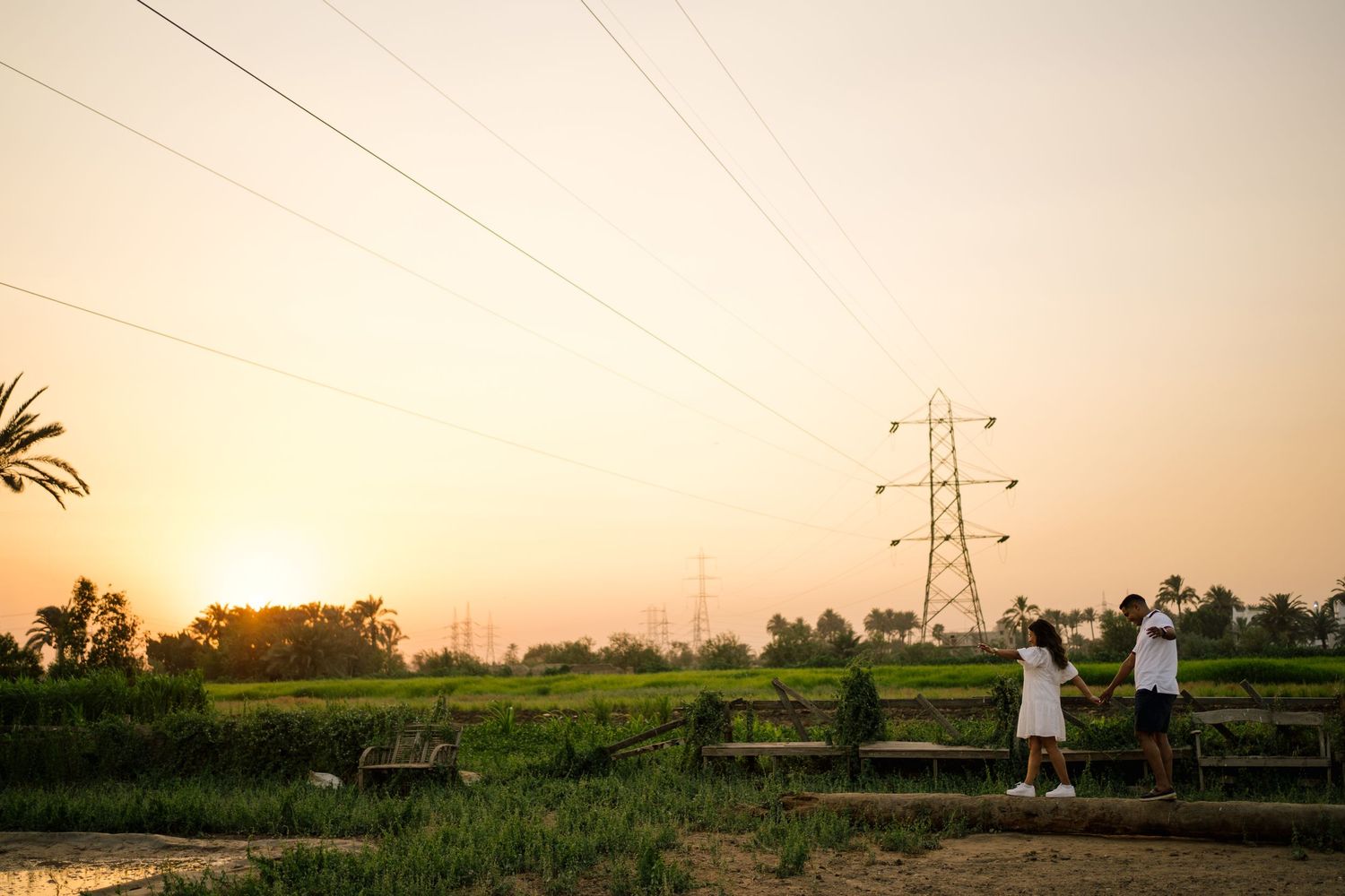 Two figures stand in a grassy field at sunset with power lines and palm trees silhouetted against the orange sky.