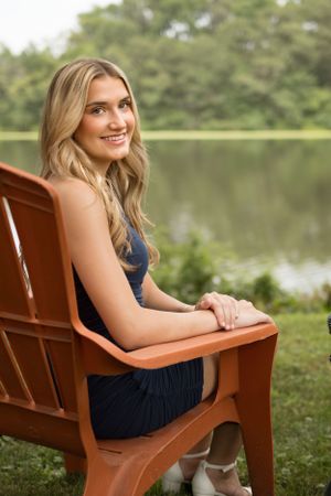 Person in a navy dress sitting on a wooden chair by a serene lake.