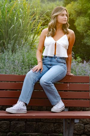 Someone sitting on a wooden bench wearing jeans and white sneakers with purple wildflowers nearby.