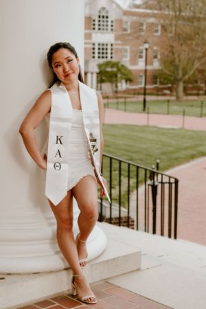 Graduate wearing a white KAO sash and dress poses on campus steps near brick buildings.
