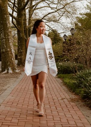Graduate in white sash and dress walks along brick pathway surrounded by trees.