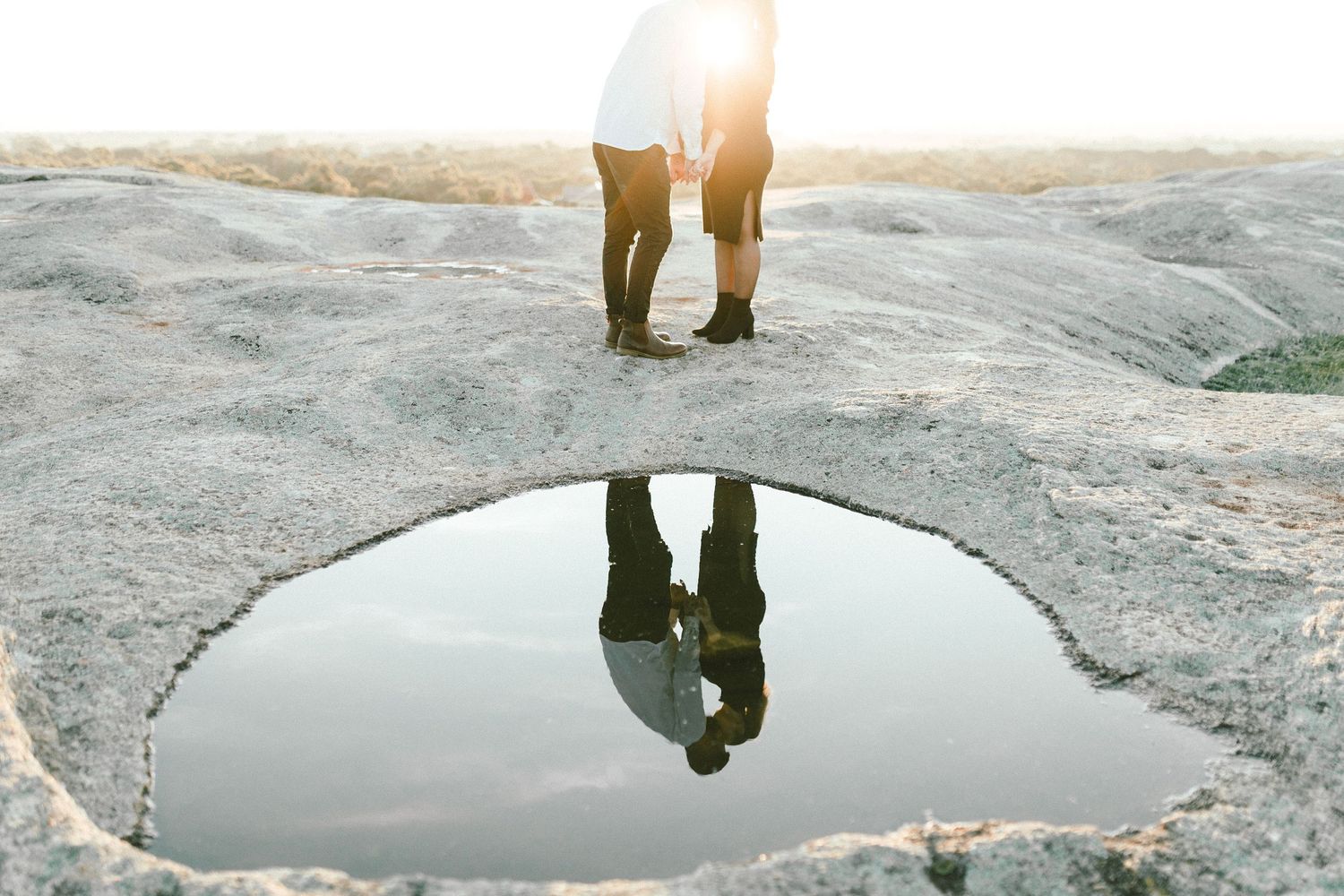 Couple's reflection appears in natural water pool on rocky surface at sunset.