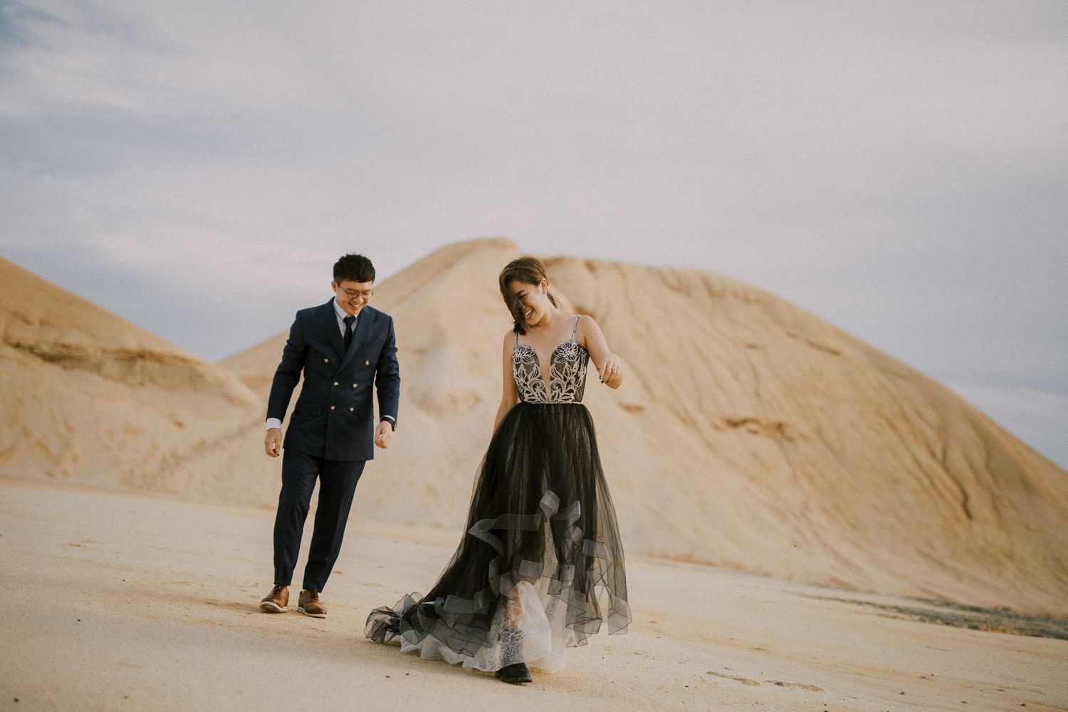 Playful couple running and sliding down sandy dunes during a desert photoshoot.