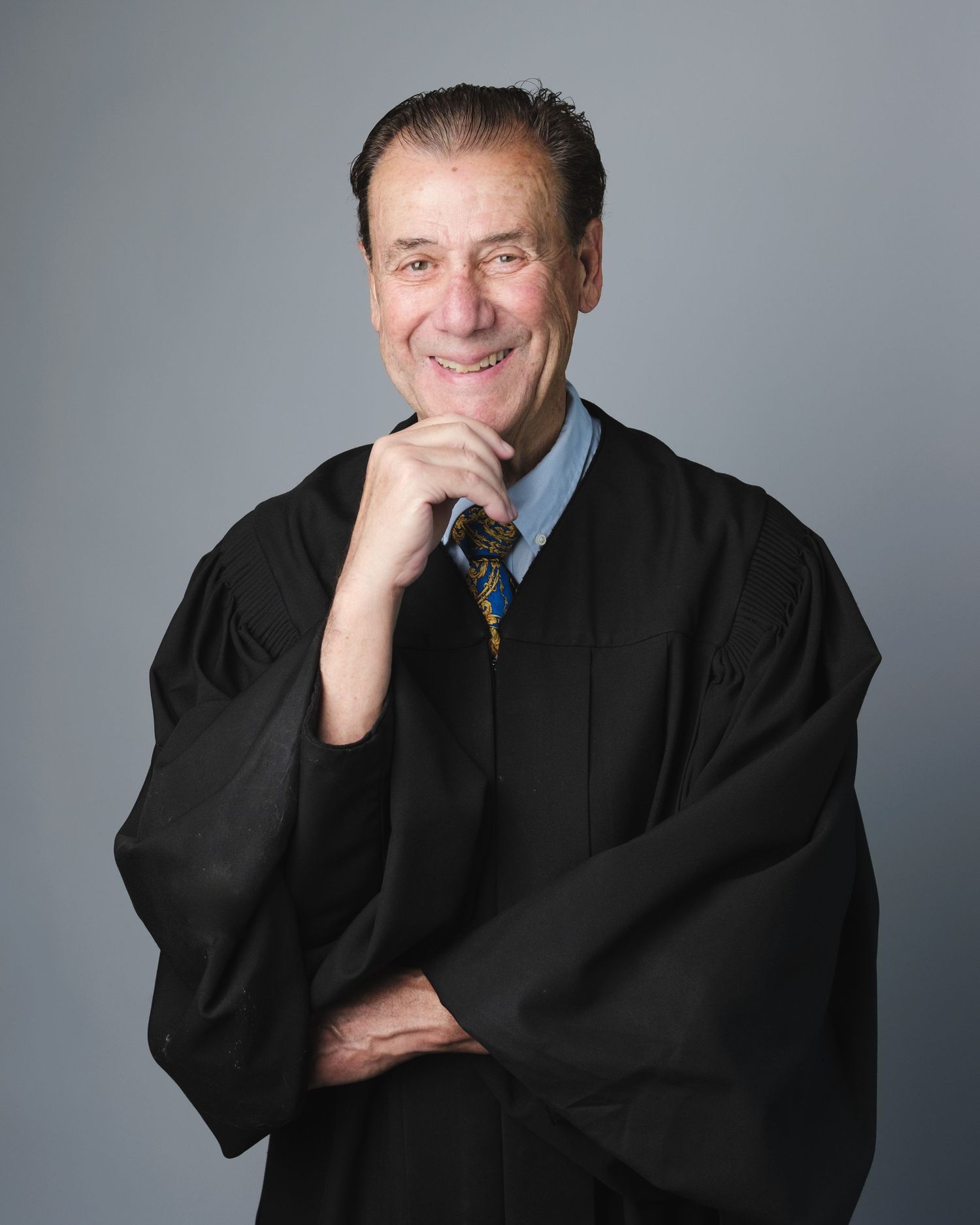 Professional headshot portrait of someone wearing a black judge's robe and blue dress shirt with tie against gray background.