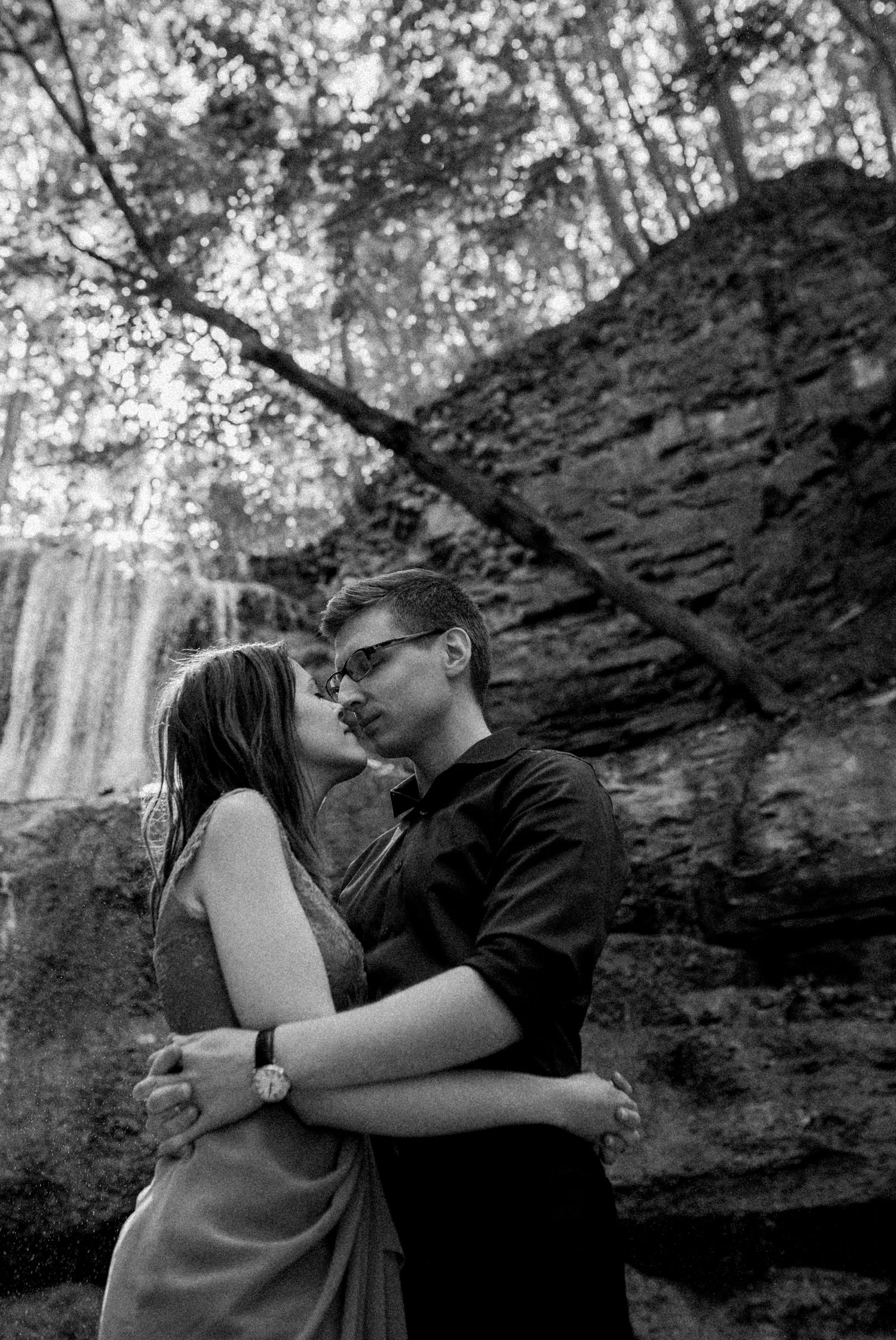 A couple lean in, holding each other as they stand outside in front of a waterfall. Photo by Katie Crane Photography.