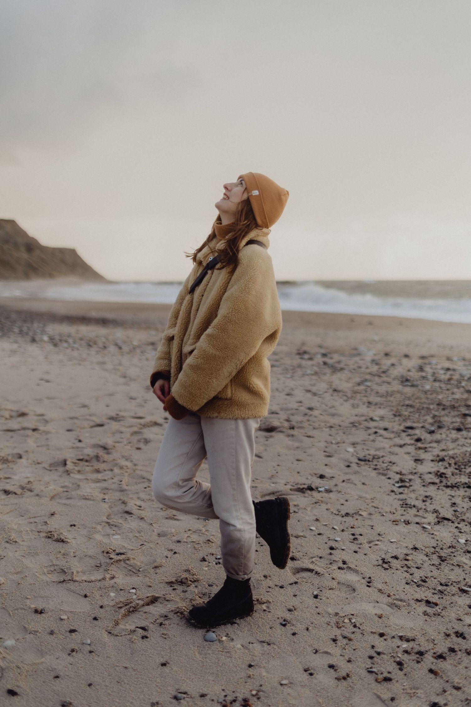 Person in beige hoodie and gray pants standing on misty beach.