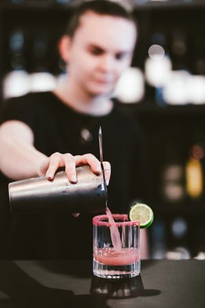 Bartender pouring a pink cocktail into a rocks glass garnished with lime.