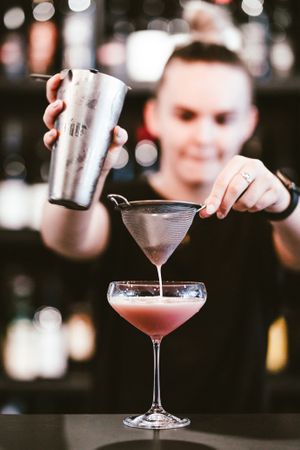 A bartender pouring a pink cocktail into a martini glass with precision.