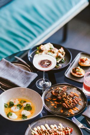 Various gourmet dishes and cocktails served on dark plates at a restaurant table.