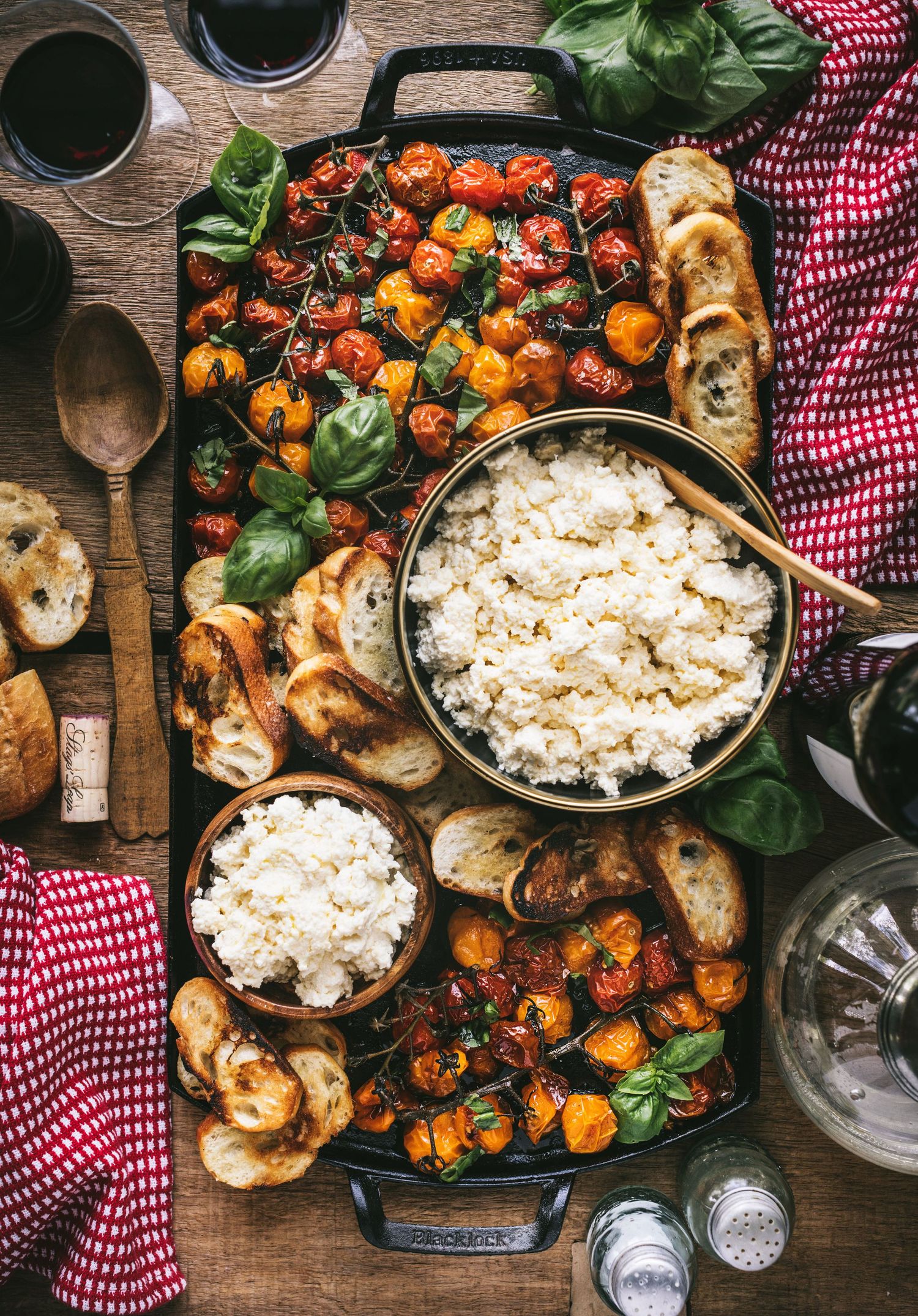 Top-down view of a rustic appetizer board with roasted tomatoes, creamy ricotta, toasted crostini, and red wine.