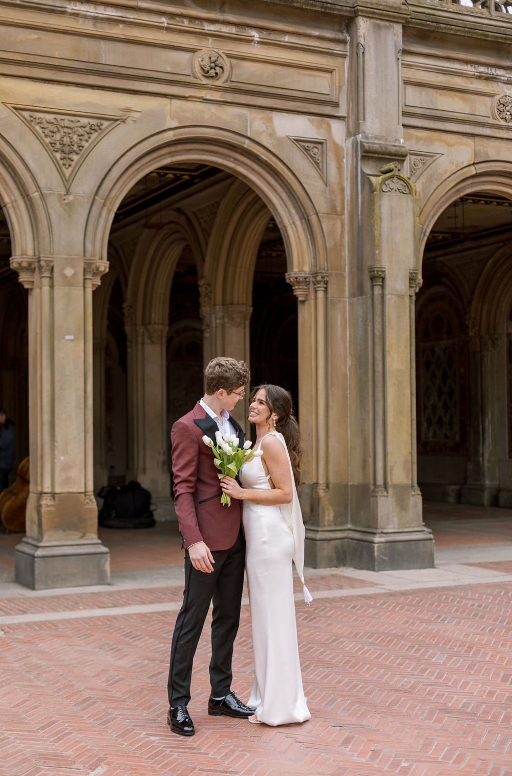 Wedding couple poses under historic stone arches with bride in sleek white dress and groom in burgundy suit.