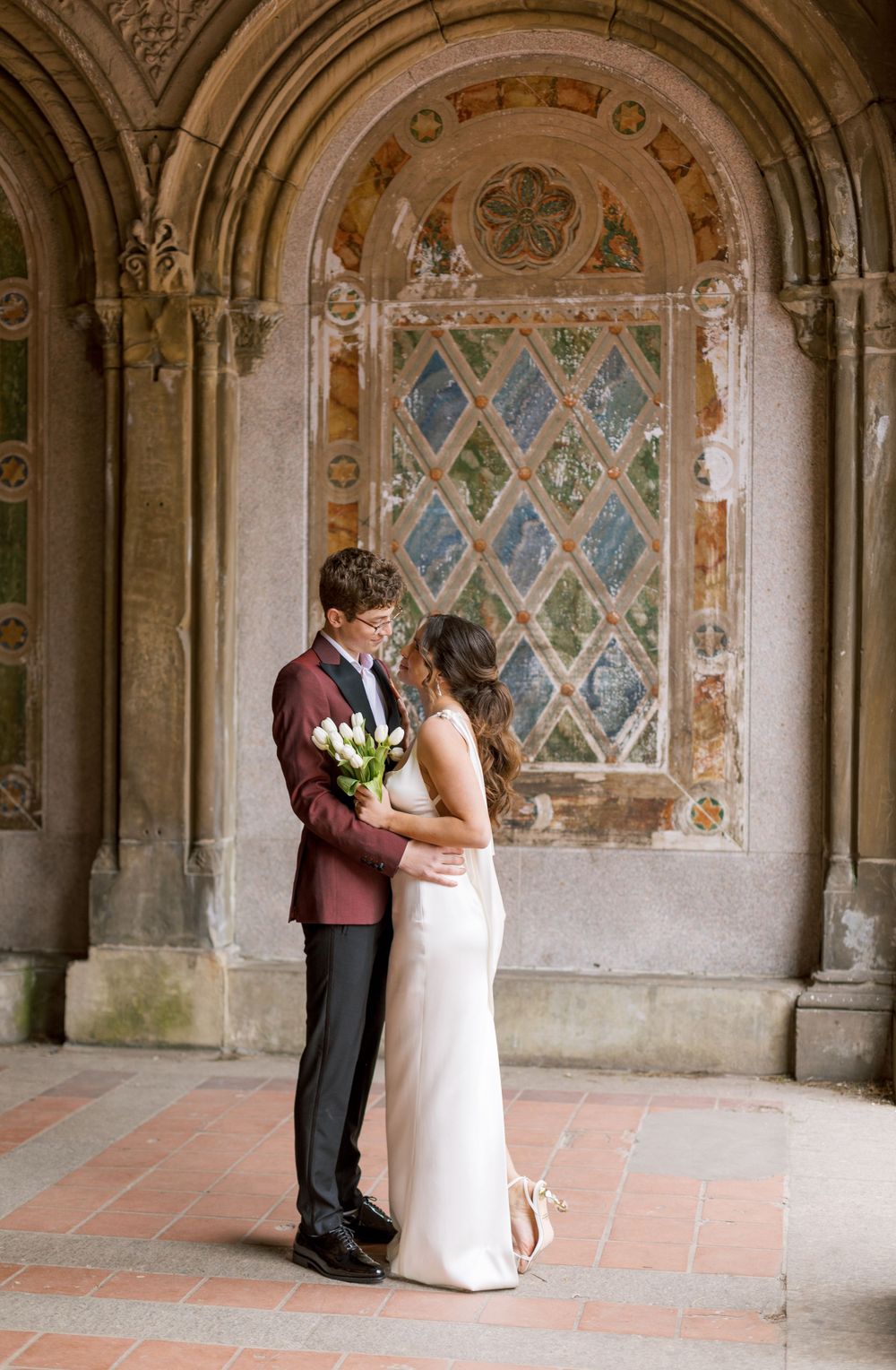 Wedding couple stands before ornate stained glass window holding white tulips.