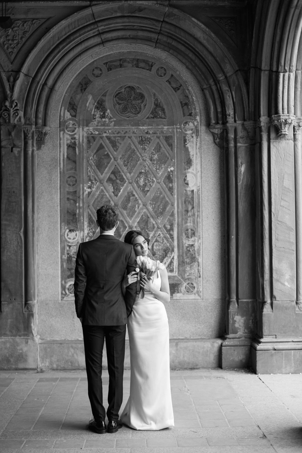Black and white wedding photo showing couple against ornate stained glass window architecture.