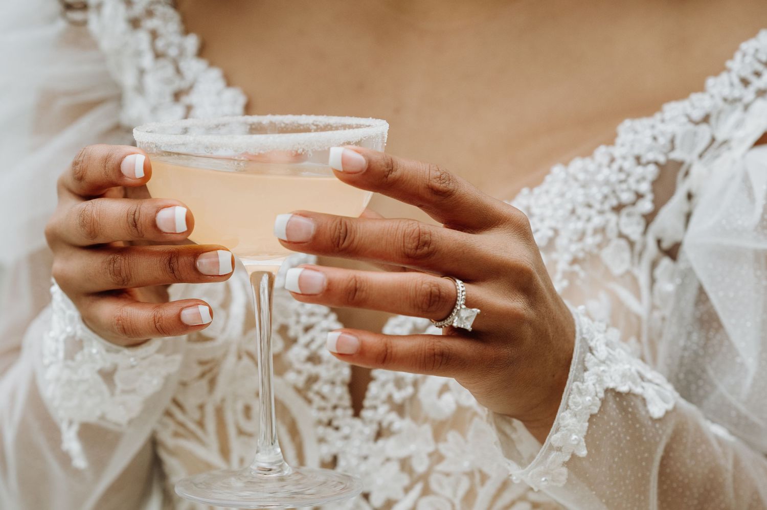 Close-up of lace wedding dress details with engagement ring and champagne glass.